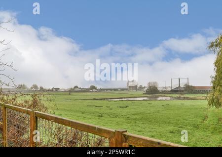 un campo verde con alberi ed edifici in lontananza, preso da dietro una recinzione di legno che si appoggia su di esso Foto Stock