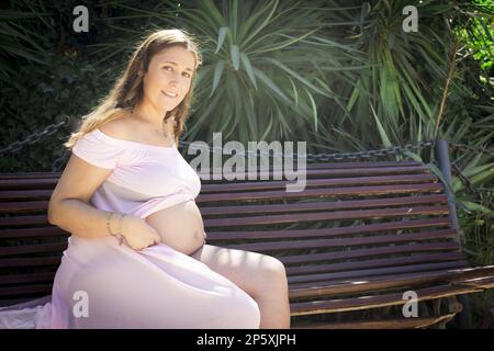 Donna incinta di sette mesi seduta su una panca di legno. Espressione molto felice Foto Stock