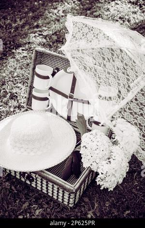 Foto in bianco e nero di un romantico set con cestino da picnic vintage, ombrellone in pizzo e cappello estivo sul prato del giardino del parco nelle giornate estive assolate all'aperto. Foto Stock