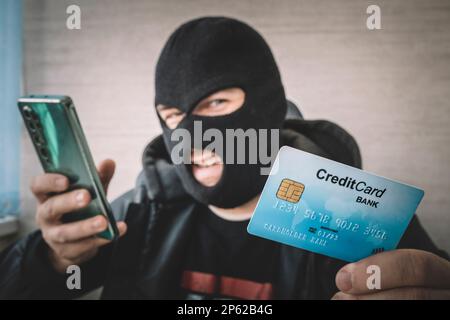 Close up di un uomo in maschera di rapina e cappa tenendo la carta di credito e guardando lo schermo dello smartphone. Penale maschio predispone un affare finanziario wit Foto Stock