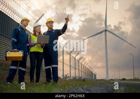 Tre tecnici tecnici in uniforme con la stazione di alimentazione della centrale di potenza della turbina eolica in piedi e di controllo. Energia pulita e ambiente. Foto Stock