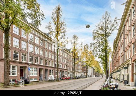 Amsterdam, Paesi Bassi - 10 aprile, 2021: Una strada cittadina con motociclette parcheggiate sul lato e alberi in mezzo alla strada, tutto lungo la strada Foto Stock