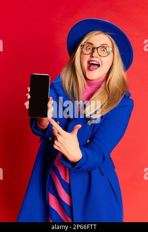 Felice donna eccitata in abiti eleganti colorati in posa emotiva, puntando lo schermo del telefono contro sfondo rosso studio Foto Stock