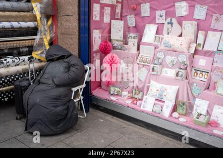 Una persona senza casa siede su una sedia nella strada principale a Cricklewood, fuori da un rivenditore che vende biglietti da visita a tema rosa, il 6th marzo 2023, a Londra, Inghilterra. Foto Stock