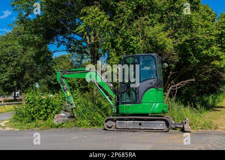 Un piccolo escavatore verde si trova sul lato della strada. Sullo sfondo di un albero verde. Foto Stock