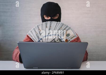 un ladro maschio tiene i dollari nella sua mano. utilizzo di un computer portatile per le attività di pirateria informatica. Concetti di criminalità informatica. Foto Stock