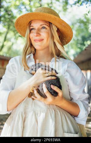 Primo piano di una giovane bella donna che tiene un piccolo coniglio nero. La bella ragazza caucasica in un cappello di paglia e una camicia bianca sorride e tiene un coniglietto carino Foto Stock