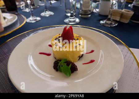 Primo piano di mousse di frutto della passione guarnita con fragole e more su un grande piatto bianco su un tavolo con una tovaglia blu e gla di cristallo Foto Stock