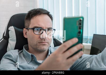 Concetto di uomo moderno di successo. Giovane ragazzo focalizzato in occhiali seduti su panca in pelle con in ufficio. Uomo d'affari caucasico millenario digitando e wa Foto Stock