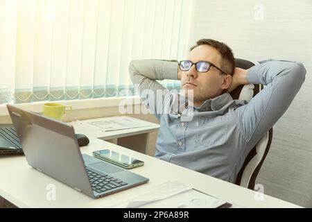 Felice rilassato caucasico giovane uomo riposo in sedia distratto dal lavoro al computer, alleviare le emozioni negative. Calmo uomo millenario in bicchieri sedersi a rilassarsi Foto Stock