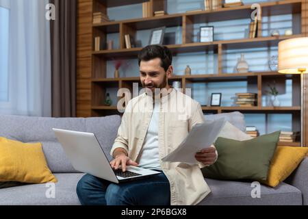 Un giovane uomo in una camicia beige si siede a casa sul divano con un computer portatile e lavora con documenti, bollette. Foto Stock