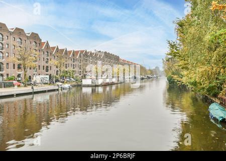 Amsterdam, Paesi Bassi - 10 aprile 2021: Alcuni edifici e barche sull'acqua di fronte a una fila di case con alberi che crescono lungo il fiume Foto Stock