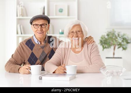 Felice coppia anziana seduta a casa con tazze di tè e sorridente alla macchina fotografica Foto Stock