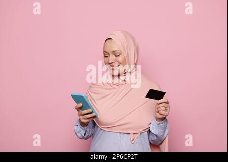 Donna musulmana in hijab rosa, utilizzando smartphone e carta di credito, effettua pagamenti online tramite app mobili e internet banking Foto Stock