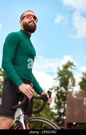 Giovane uomo sportivo sorridente in occhiali con barba in piedi con bici nel parco estivo, allenamento cardio in bicicletta, passeggiate in città in bicicletta, controllo del peso Foto Stock