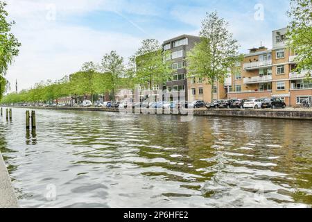Amsterdam, Paesi Bassi - 10 aprile, 2021: Alcuni edifici sul lato di un corpo d'acqua con auto parcheggiate lungo di esso e alberi nelle fortune Foto Stock