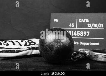 Palla di metallo con catena, uniforme di prigione e cartoncino da lettera mugshot su tavolo di legno nero Foto Stock