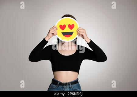 Donna che copre il viso con cuore occhi emoji su sfondo grigio Foto Stock