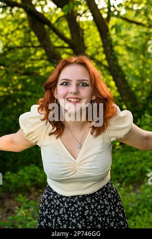 Una giovane donna ridente dai capelli rossi, che indossa una blusa di colore chiaro, braccia tese di lato, di natura verde Foto Stock