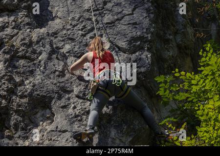 Una giovane donna con una corda impegnata arrampicata sulla scogliera. In estate, una donna sale su una parete rocciosa verticale in maglia rossa Foto Stock