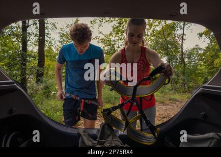 Giovane arrampicatore maschio e femmina che prende l'attrezzatura di arrampicata dal bagagliaio dell'auto e mette le imbracature sopra e si prepara per l'avventura Foto Stock