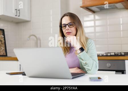 Donna bianca adulta in occhiali che legge il testo dallo schermo del computer. Donna Ucraina concentrata e penosa che lavora sul computer portatile a casa Foto Stock