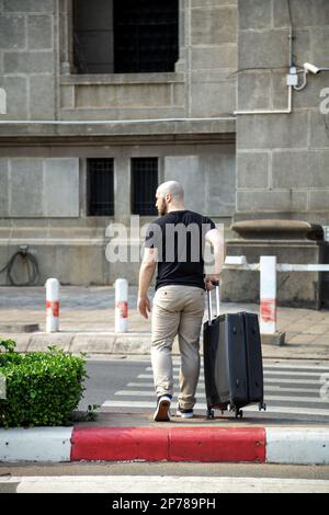 Un giovane uomo in abiti casual che attraversano la strada, tenendo una valigia visto da dietro. Un ragazzo calvo in un viaggio d'affari che arriva all'aeroporto a piedi Foto Stock