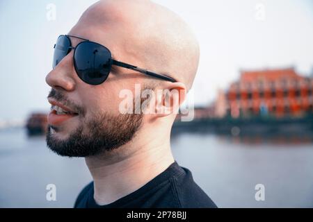 Felice giovane uomo in occhiali da sole che passeggiano lungo un fiume e sognando. Ragazzo maschile calvo che sorride da vicino Foto Stock