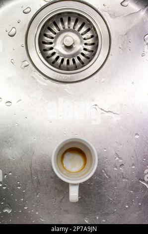 Piccola tazza di caffè sporca in porcellana bianca in un lavandino in metallo accanto al foro di scarico vista dall'alto. Foto Stock