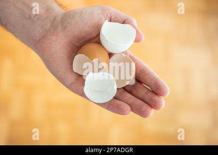 Gusci di pollo spezzati e aperti tenuti in mano dall'uomo. Primo piano, vista dall'alto, sfondo marrone sfocato. Foto Stock