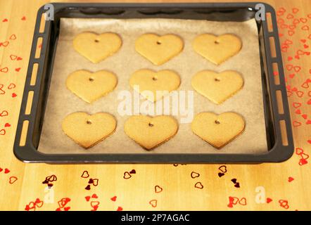 Biscotti romantici al cuore su una teglia da forno sul tavolo con molte decorazioni a cuore rosso. Foto Stock