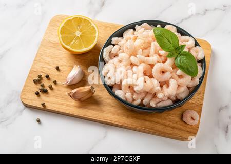 Gamberetti surgelati bolliti in una ciotola sopra il tagliere. Code di gamberi sbucciate cotte, limone e aglio preparati per la cottura. Ricetta di pesce a basso contenuto calorico. Foto Stock