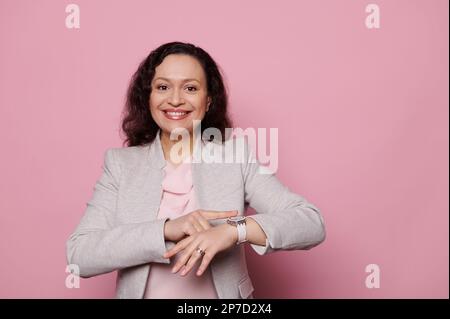 Donna matura sorridente che controlla il tempo sull'orologio da polso, guardando la macchina fotografica, isolata su sfondo rosa. Puntualità. Persone Foto Stock