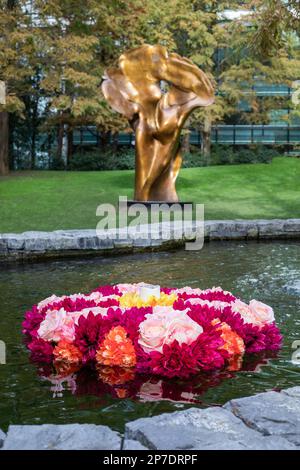 Fiori di rangoli colorati galleggiano in una piscina d'acqua di fronte all'opera d'arte in bronzo intitolata Fortuna di Helaine Blumenfeld nel Jubilee Park, Canary Wharf Foto Stock