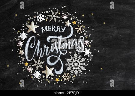 Buon Natale scritto in gesso su una lavagna nera con confetti e decorazioni natalizie, piatto disteso visto dall'alto Foto Stock