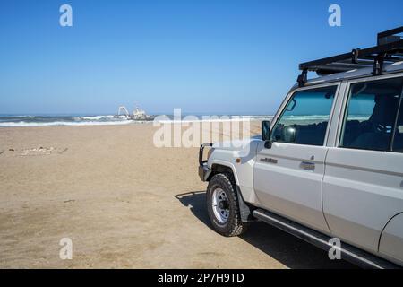 Il LandCruiser si trova sulla spiaggia in vista di un naufragio danneggiato in mare. Swakopmund, Namibia, Africa Foto Stock