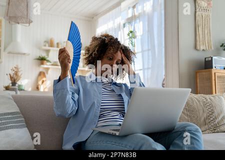 Donna nera soffre di colpo di calore piano senza aria condizionata ondeggiante ventilatore seduto sul divano a casa Foto Stock