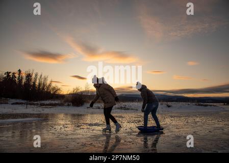 Due donne pattinano sul ghiaccio su un lago selvaggio nel Canada settentrionale all'inizio della stagione invernale con uno splendido sfondo arancione al tramonto. Foto Stock