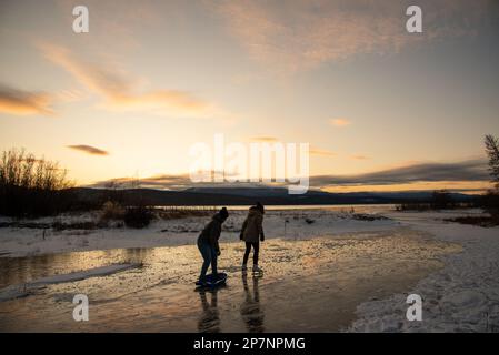 Due donne pattinano sul ghiaccio su un lago selvaggio nel Canada settentrionale all'inizio della stagione invernale con uno splendido sfondo arancione al tramonto. Foto Stock