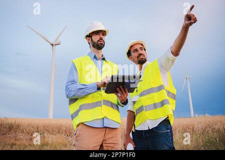 Due ingegneri di turbine a vento che lavorano insieme, un tecnico che utilizza un dispositivo tablet per vedere l'efficienza del suo caposquadra indicando quale generatore deve essere riparato. Concetto di energia rinnovabile. Foto di alta qualità Foto Stock