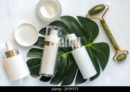 Set di cosmetici e rullo massaggiatore in giada per la cura della pelle con foglia verde su un tavolo in marmo. Concetto di prodotti per la cura della pelle Foto Stock