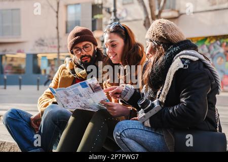 Giovane gruppo di amici turisti persi in cerca di posizione su una mappa seduta sulla strada. Tre amici che leggono una guida per trovare un monumento in un viaggio in città. Concetto di viaggio. Foto di alta qualità Foto Stock