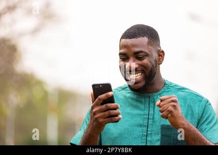 Excited Young African Man Holding smartphone all'aperto, felice di vincere la lotteria. Foto Stock