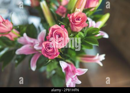 Un bel bouquet di rose rosa e corallo e lillies in un vaso chiaro caraffa su un tavolo da pranzo in legno scuro con luce naturale attraverso le finestre Foto Stock