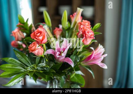 Un bel bouquet di rose rosa e corallo e lillies di giglio in una caraffa di vaso chiaro su un tavolo da pranzo in legno scuro con luce naturale che viene attraverso Foto Stock