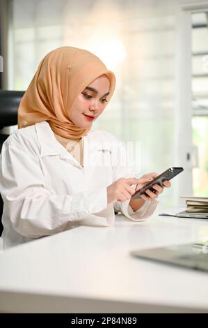 Ritratto, attraente donna d'affari musulmana asiatica millennial che indossa hijab utilizzando il suo smartphone alla sua scrivania ufficio. Foto Stock