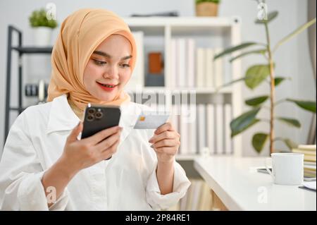 Bella donna musulmana asiatica millenaria che indossa hijab utilizzando l'app mobile banking per pagare le bollette dello shopping online, tenendo il telefono e la carta di credito. Foto Stock