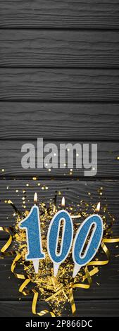 Numero 100 candela blu da celebrazione e coriandoli d'oro su fondo di legno scuro. 100th biglietto di compleanno. Concetto di anniversario e compleanno. Banner verticale Foto Stock