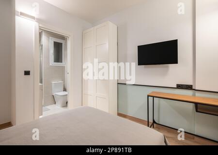 Letto matrimoniale con biancheria grigia, armadio a muro con biancheria bianca e TV con affaccio su un bagno compatto con servizi igienici e finestra. Concetto di un piccolo ed elegante appartamento Foto Stock