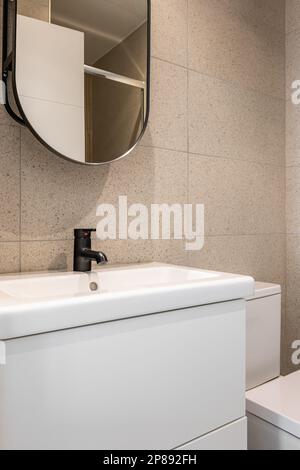Frammento di specchio e lavello con rubinetto nero e water ciotola in bagno su sfondo di piastrelle beige. Concetto di un bagno semplice ma conciso Foto Stock